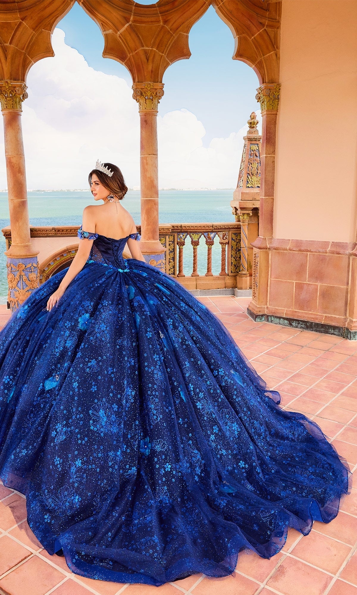 A woman in the Quinceanera Dress PR30159 By Princesa, a blue off-shoulder gown with a corset bodice and tiara, stands on a tiled terrace with ornate arches overlooking a body of water.
